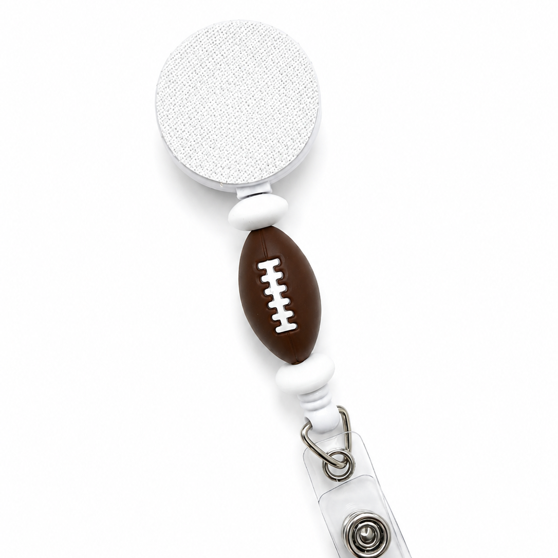 Football badge reel featuring a brown football focal bead with white football laces and white accent beads on a white retractable badge reel. Cute sports-themed work badge accessory.