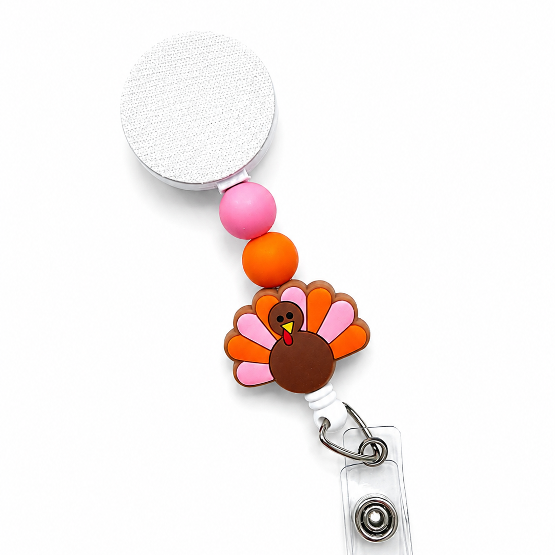 Turkey badge reel featuring a cute brown turkey with pink and orange feathers, pink and orange decorative beads, and a white retractable badge reel. Festive Thanksgiving work badge accessory.