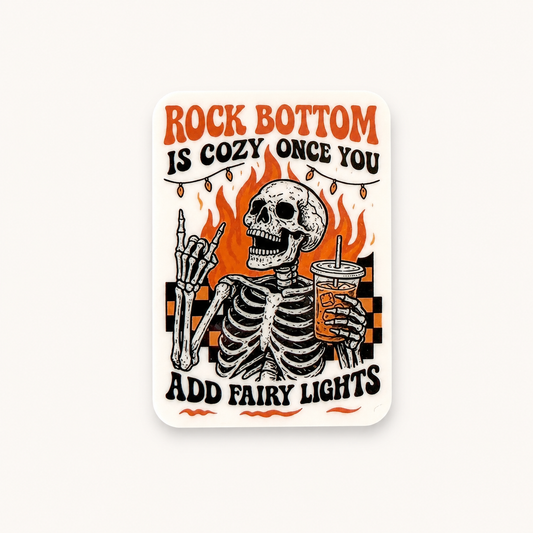 Funny skeleton sticker with “Rock Bottom Is Cozy Once You Add Fairy Lights” quote, retro spooky aesthetic with dark humor design.