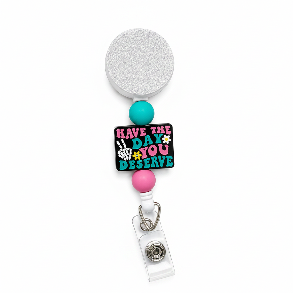 Have The Day You Deserve badge reel featuring a black statement topper with pink and teal lettering, skeleton hand peace sign, daisy accents, and teal and pink decorative beads on a white retractable badge reel.