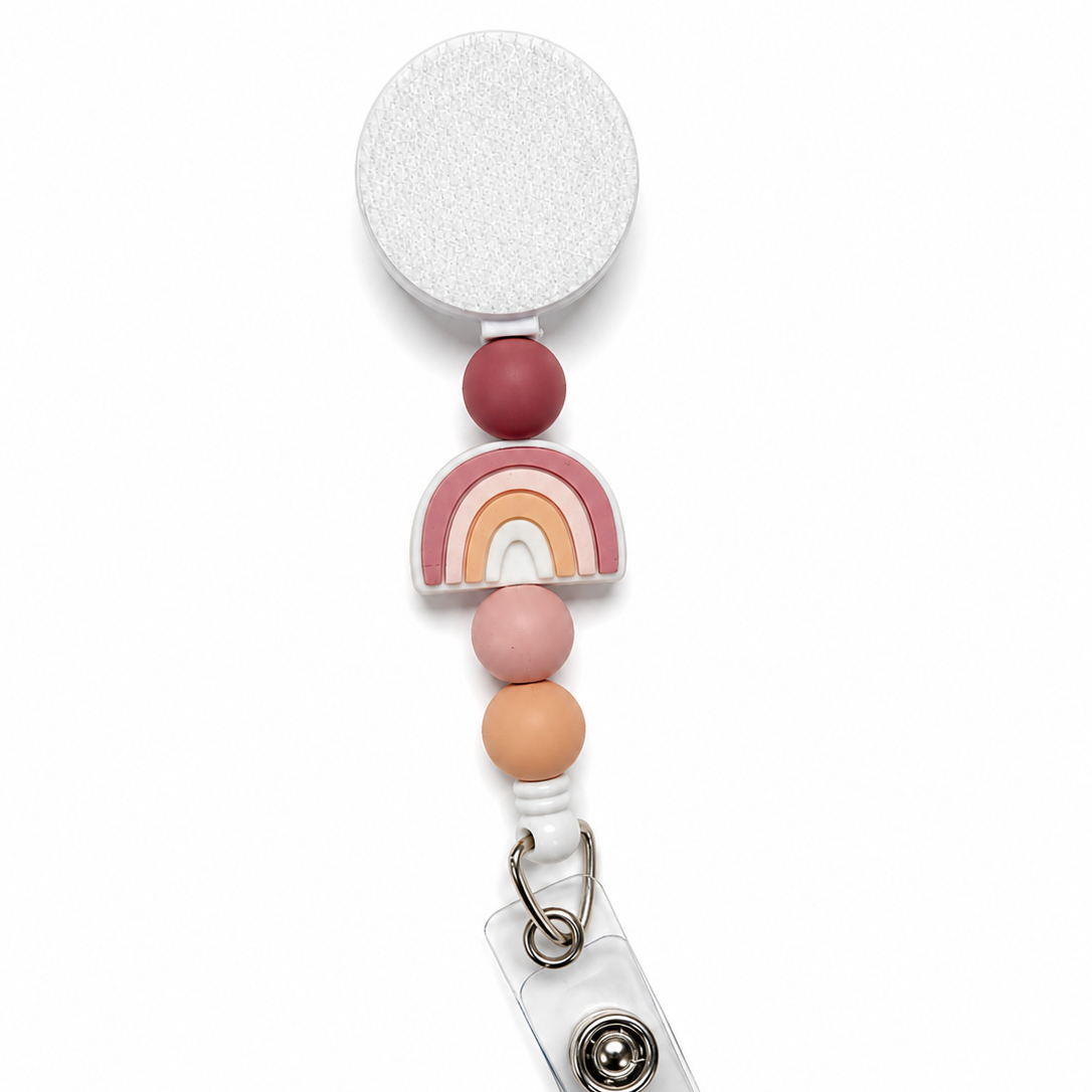 Modern Rainbow badge reel featuring a pastel rainbow centerpiece with mauve, blush pink, and peach decorative beads on a white retractable badge reel. Boho work badge accessory.
