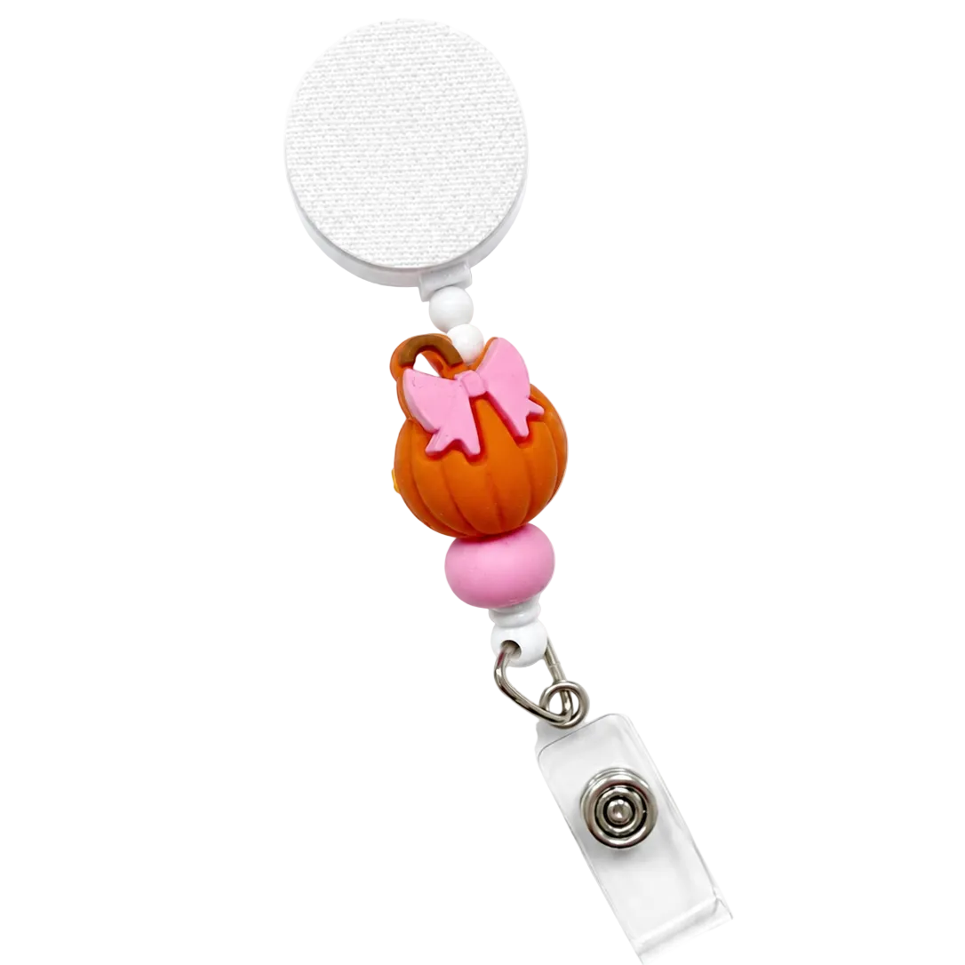 Pink Bow Pumpkin badge reel featuring an orange pumpkin with a pink bow, pink decorative bead accents, retractable badge reel, and secure badge holder attachment for Halloween workwear.